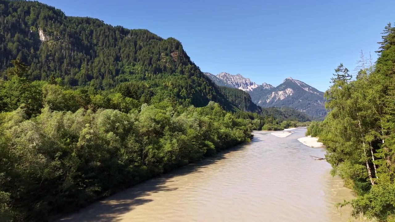 Droneutsikt over den gjørmete elven Lech som renner langs et bratt, skogkledt fjell med Alpene i forgrunnen, Füssen, Tyskland.