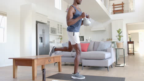 hombre afroamericano concentrado haciendo ejercicio con kettlebell en la sala de estar soleada, cámara lenta