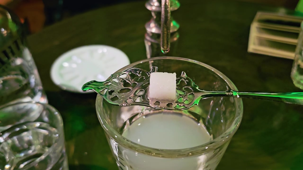 Toma macro de un terrón de azúcar sobre una cuchara de absenta ornamentada encima de un vaso, mientras agua fría gotea de una fuente, creando un efecto louche turbio en un salón de cócteles verde tenue.