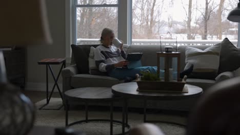 woman sitting on the couch in front of a bay window, drinking coffee while scrolling on a tablet from far away