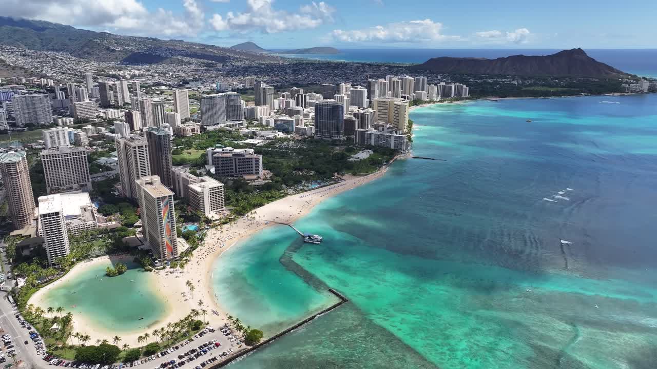 Aerial drone footage over Waikiki Beach in Honolulu, Oahu, Hawaii, showcasing turquoise ocean water, palm trees, hotels, surfers, and the tropical coastline of this world-famous vacation destination
