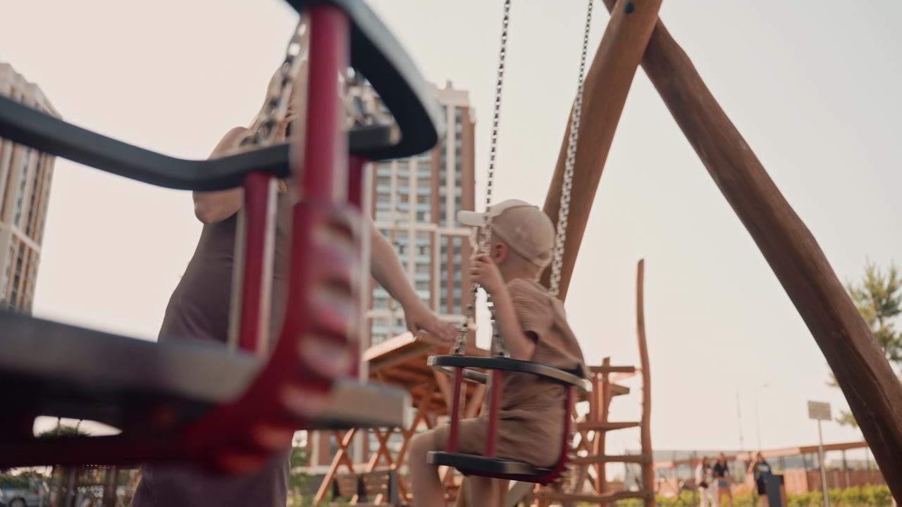 Una donna parla al telefono mentre un bambino gioca sull'altalena al parco giochi. La scena si svolge all'aperto con alti edifici sullo sfondo, caratterizzata da luce naturale e ombre tenui.