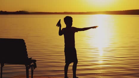 boy throws paper airplane. silhouette of a boy on an old bridge by the river. beautiful summer sunset