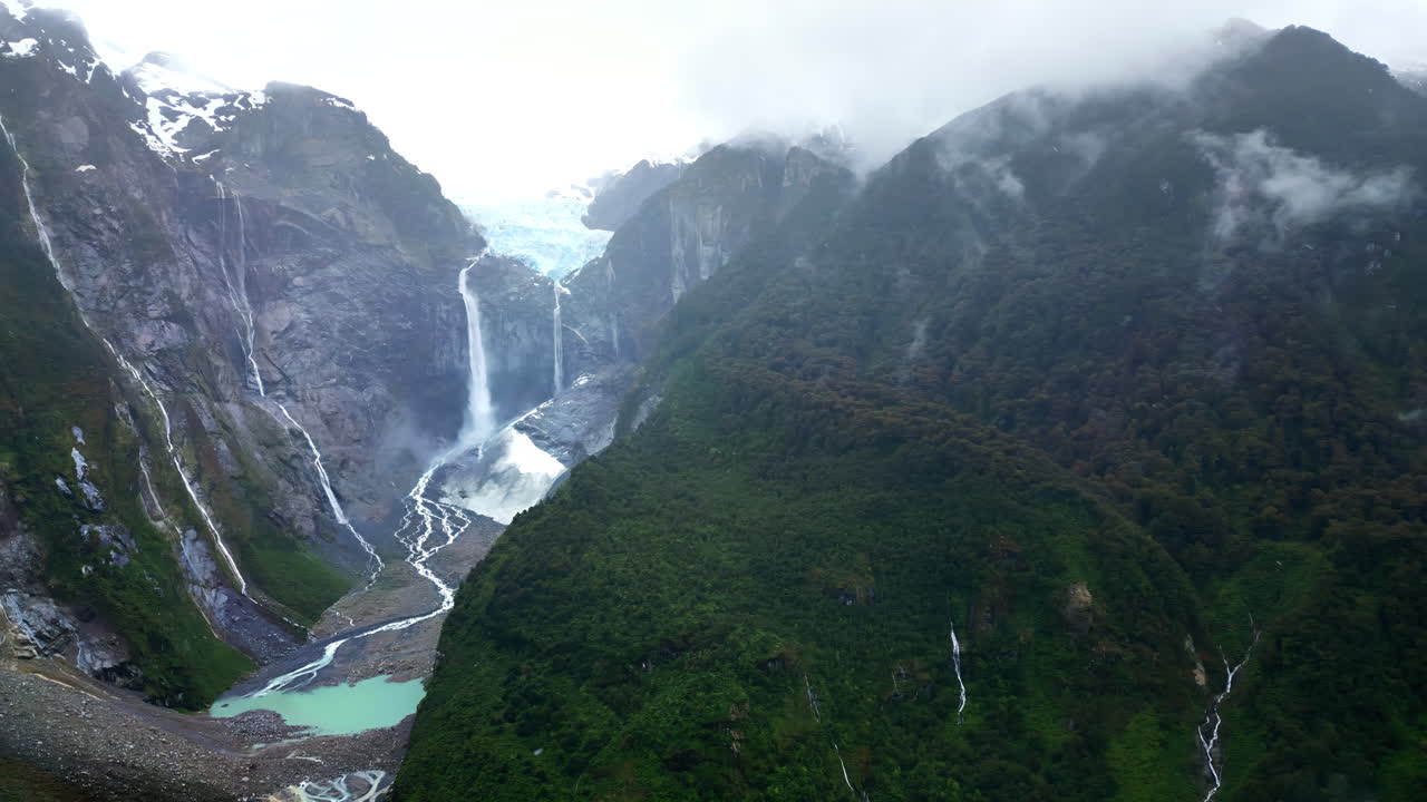 Aerial drone view of the stunning Ventisquero Colgante glacier waterfall cascading down steep cliffs surrounded by lush green forest in slow motion