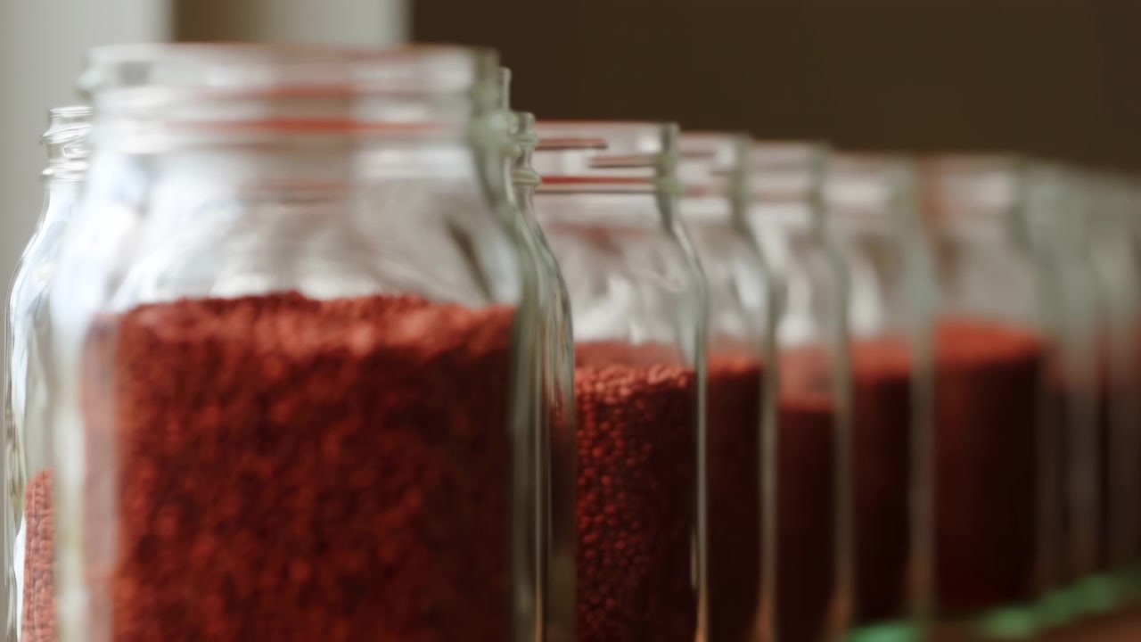 A clear glass jar packed with red lentils stands in the foreground, with several similar jars softly blurred behind it. Warm natural light emphasizes the