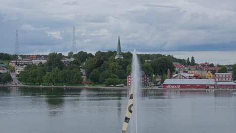 Water-fountain-on-the-shore-city-of-motala,-red-swedish-house,-drone-telephoto