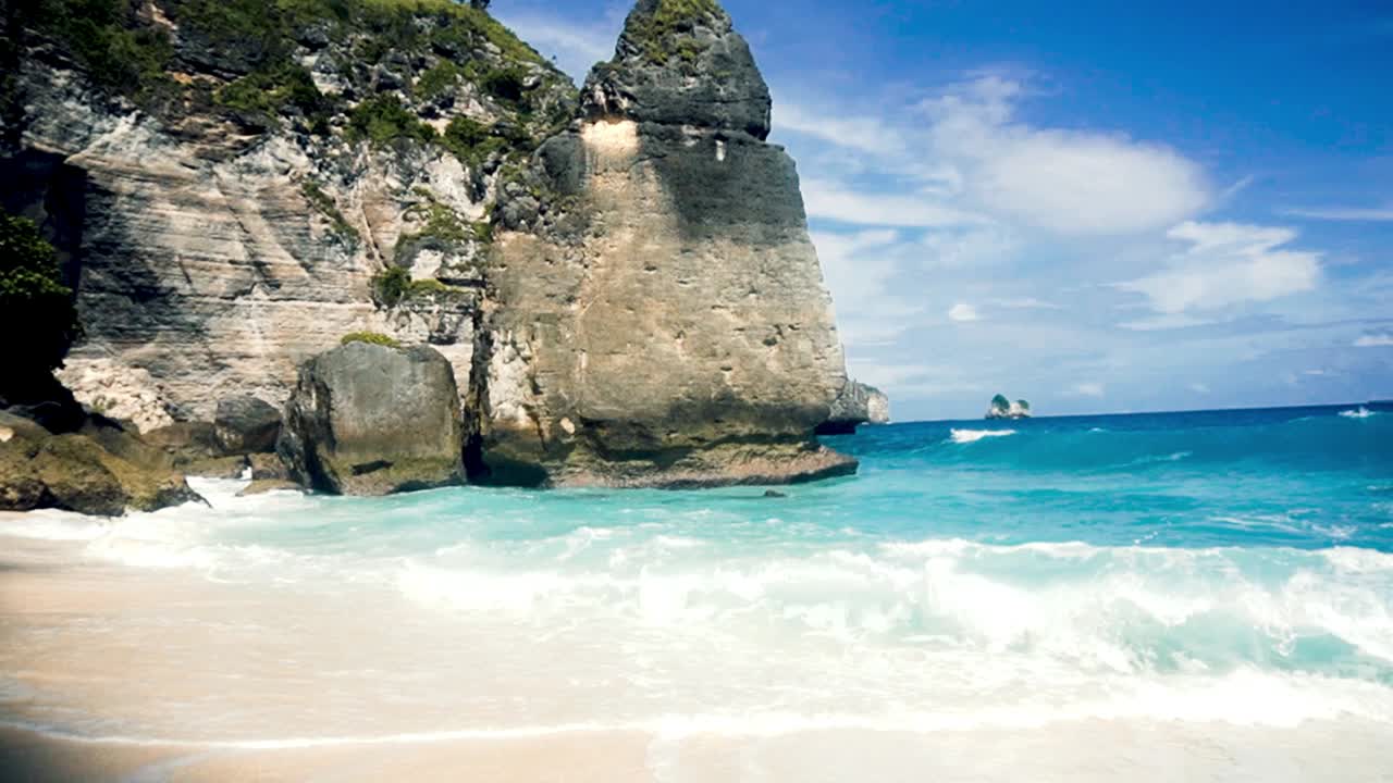 Static shot of waves crashing on a white sand clear water beach in Bali, Indonesia, during a sunny day