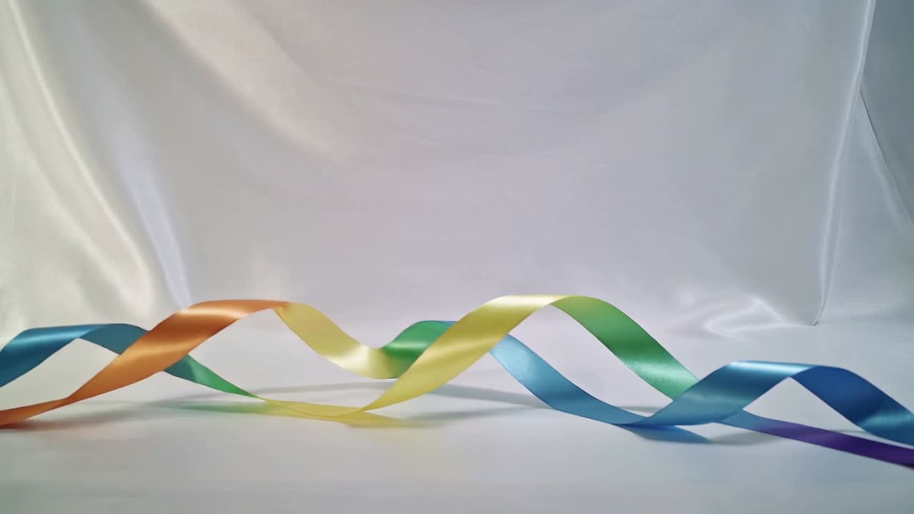 Al inicio, cinta de satén multicolor cambiante formando bucles sobre una mesa blanca, mostrando colores giratorios.