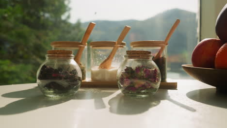 herbal tea ingredients put on table closeup. glass jars with dried flowers, herbs and sugar on white countertop by window. set for organic beverage