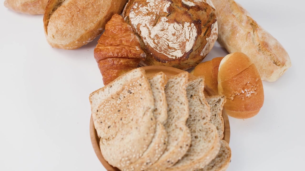 Ripresa aerea a rotazione lenta di pane a lievitazione naturale, croissant e pane integrale a fette su una superficie bianca.