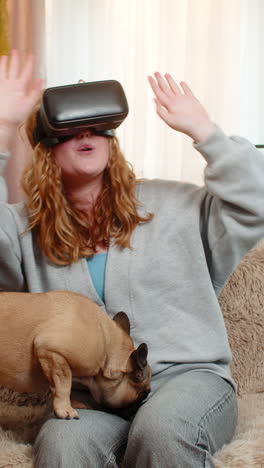 Young woman using vr headset on home sofa with excited movement as pug dog watches with funny face