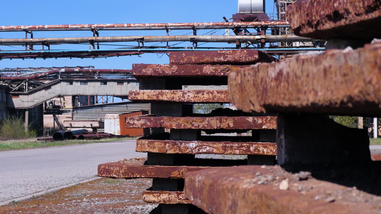 Exterior view of abandoned Soviet heavy metallurgy melting factory Liepajas Metalurgs territory, rust-covered metal scrap piles, sunny day, heat pipelines in background, medium shot