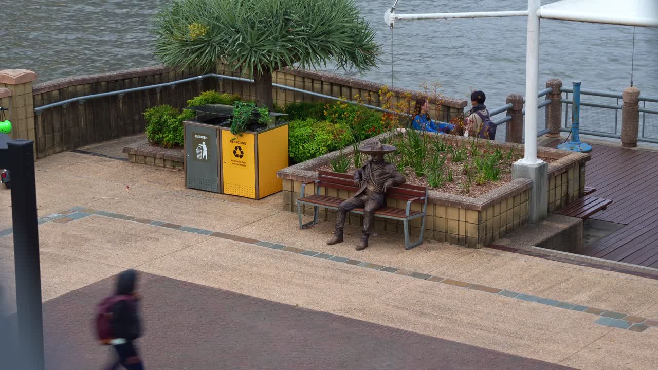 A living statue spotted sitting on the park bench by a riverside promenade in the south bank parklands, Brisbane city, time-lapse fast motion shot.