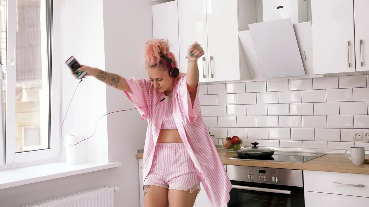 Giovane donna felice con i capelli rosa che balla in cucina, indossando vestiti rosa e ascoltando musica con le cuffie