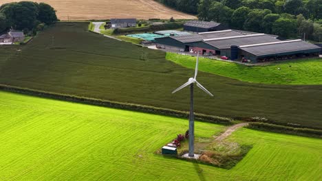 aerial view of a wind turbine spinning in vibrant green farmland near agricultural buildings, captured in daylight with smooth camera movement over kinross, scotland