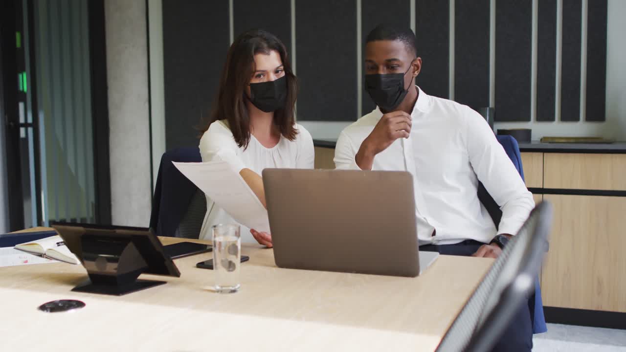 Diverse businessman and businesswoman wearing face masks discussing and using laptop in office