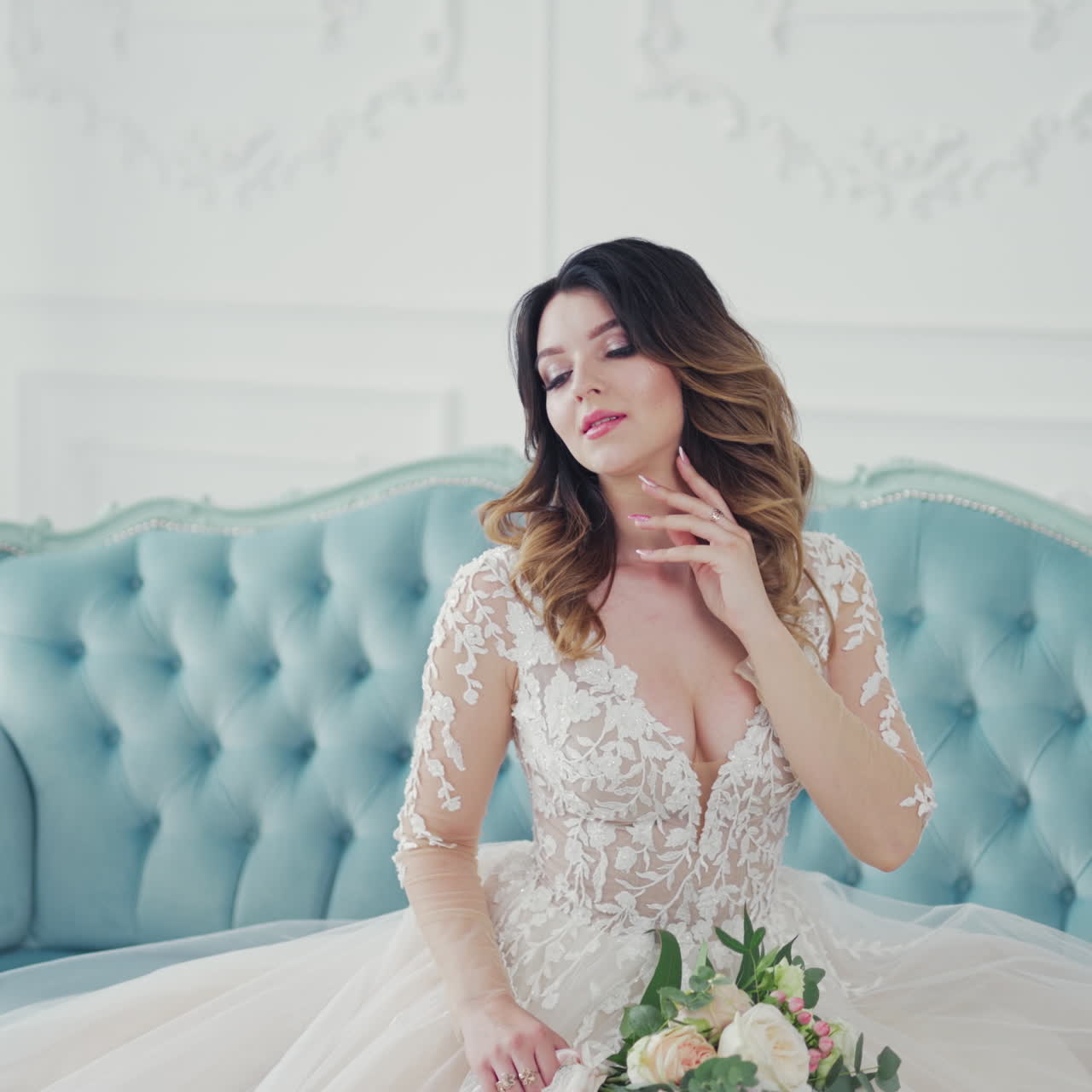 Gorgeous young woman on the couch with her hand on neck. Luxurious bride in a wedding dress with bouquet of flowers is waiting for the groom at home.
