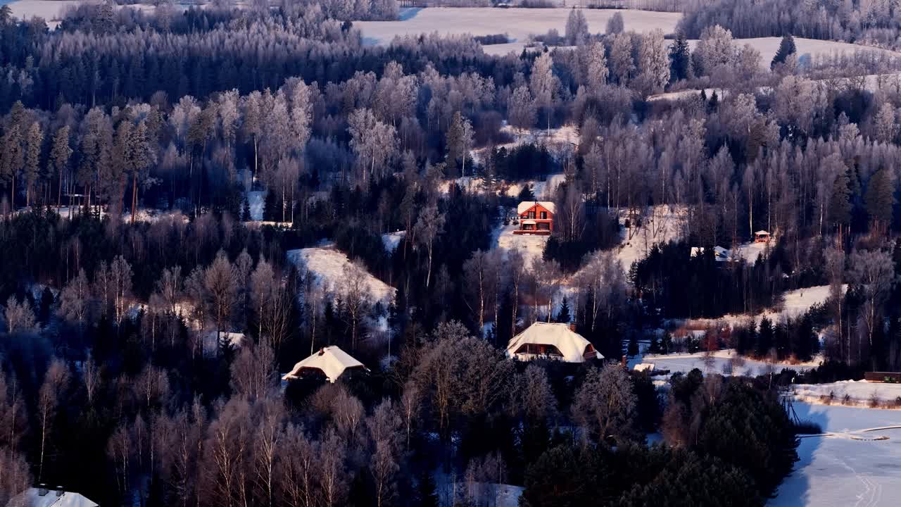 Talvinen dronekuva puumökistä, joka erottuu lumipeitteisten kukkuloiden ja puiden keskeltä. Kylmät siniset sävyt kontrastoivat lämpimien puunvärien kanssa, luoden rauhallisen talvimaiseman vahvalla polttopisteellä.