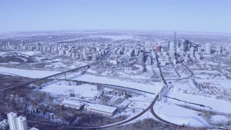 super high aerial pan out of winter snow covered city with gradaitions blue clear sky of a curvy horizon with bridges connecting to the downtown core crossing icy snow covered rivers parks forests