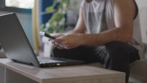 joven hombre adulto trabajando desde casa en su sala de estar usando una computadora portátil, distraído del trabajo con un teléfono nuevo correo aparece, sentado en el sofá con ropa casual