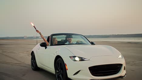 a blonde girl holds hand fire above her head and drive with her bearded guy in a blue shirt on a white convertible car against the river and a gray sky