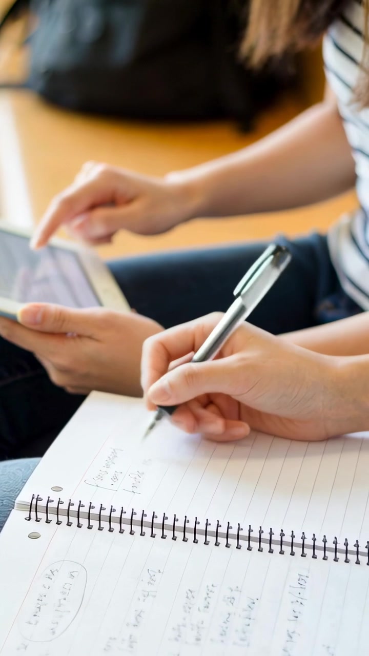 Vertical video: Tapping tablet, two students taking notes on bench with spiral notebook and pen