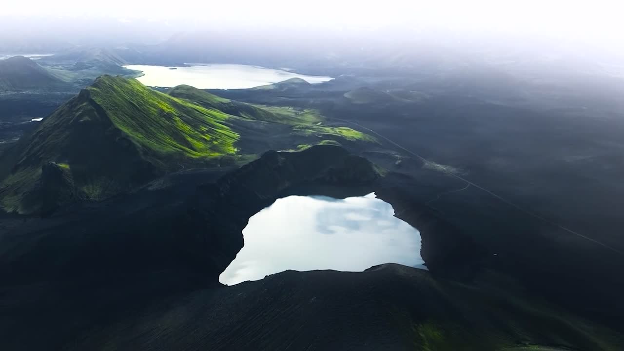 Aerial drone footage of a lake and green moss and grass covered steep tall mountains during a cloudy day in Greenland or Iceland. The lakes are reflective and white during cloudy day with mist and fog