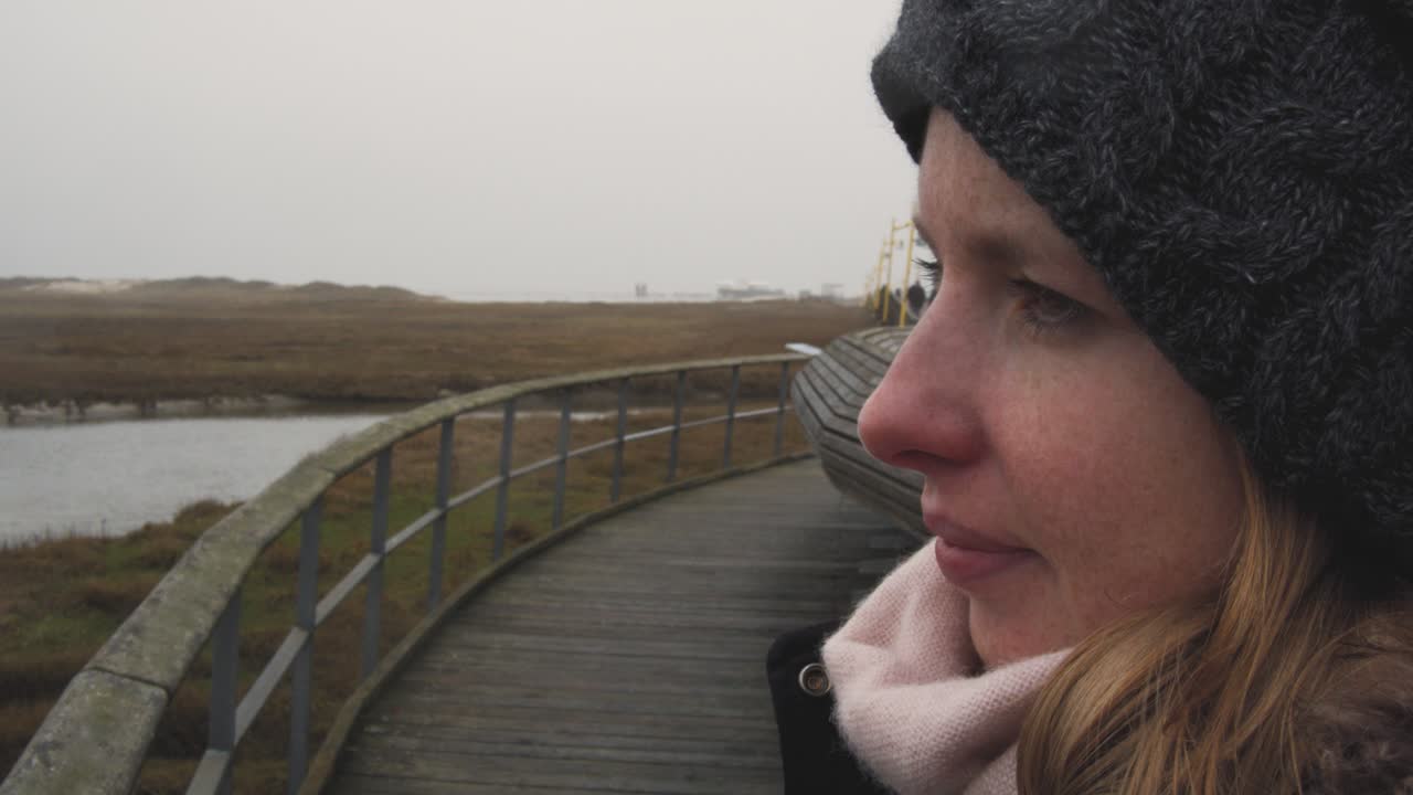 Young woman stands on a jetty and overlooks a dune landscape in the freezing cold