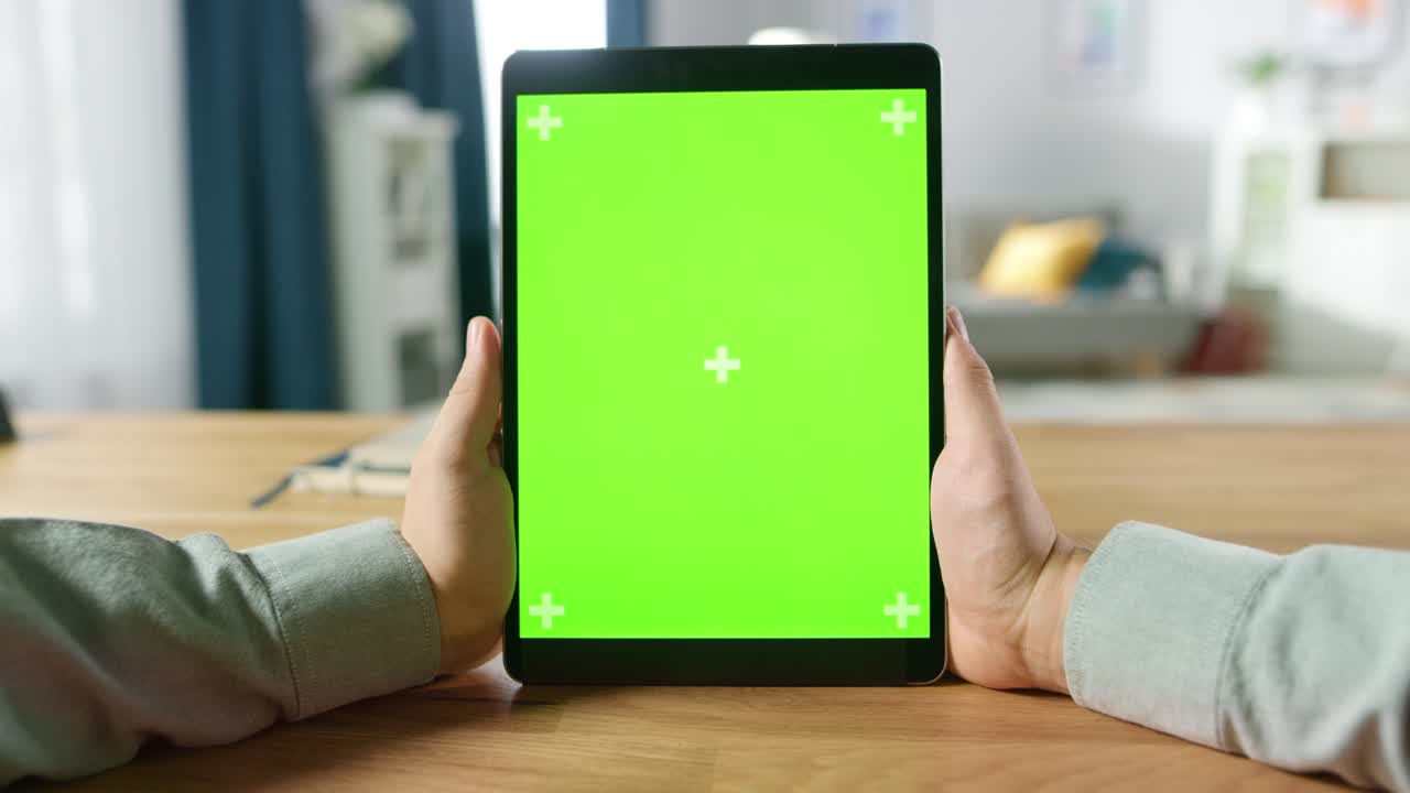 Førstepersonsperspektiv af mand der bruger grøn mock-up skærm digital tablet computer i portrættilstand, mens han sidder ved sit skrivebord. I baggrunden hyggelig stue
