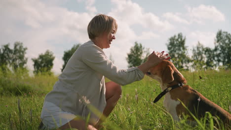 mujer alimentando al perro en un vasto campo de hierba mientras se agacha en un día soleado, compartiendo un vínculo afectuoso con su perro mirando con amor, rodeada de naturaleza verde exuberante y árboles