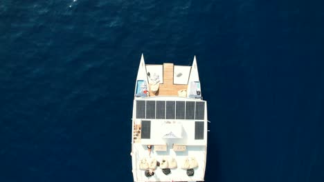Luftaufnahme-Von-Oben-Nach-Unten-Auf-Eine-Moderne-Katamaran-Yacht,-Die-Im-Blauen-Meer-Kreuzt,-Und-Ein-Paar,-Das-Sich-Auf-Dem-Dach-Sonnt
