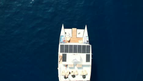 Vista-Aérea-De-Arriba-Hacia-Abajo-De-Un-Moderno-Catamarán-Navegando-En-El-Mar-Azul-Y-Una-Pareja-Tomando-El-Sol-En-La-Azotea