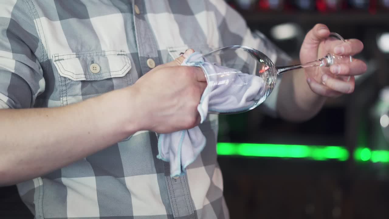 Male barman is wiping a wet glass with dry napkin on the bar background. Bartender polishes a glass near the counter