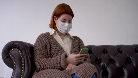 cuarentena durante la pandemia de coronavirus. mujer usando el teléfono en casa, usando una máscara médica, sentada en un sofá.