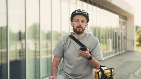 confused food delivery guy wearing thermal backpack takes a look at his smartphone to know where to go with his bike