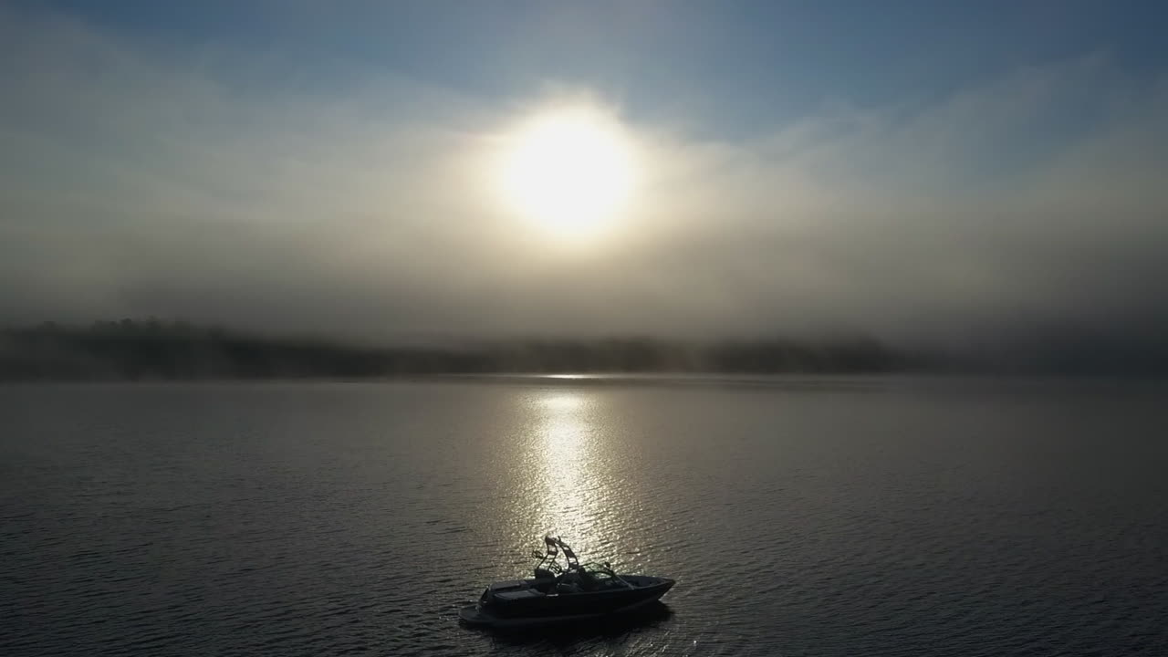 안개가 자욱한 아침 공중 스키 보트가 금색 태양 광선에 있는 호수 표면에 떠 있습니다.