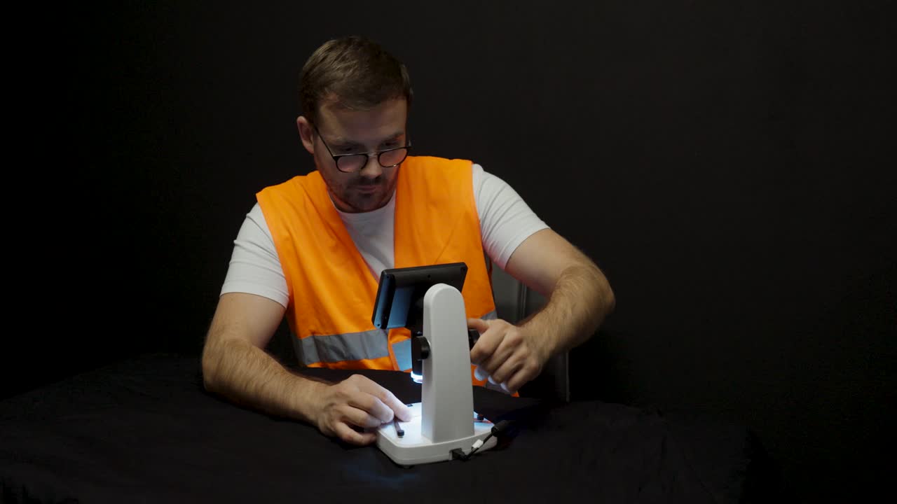 A technician in a safety vest sits at a lab table and operates a digital microscope carefully, focusing on precision and workplace safety