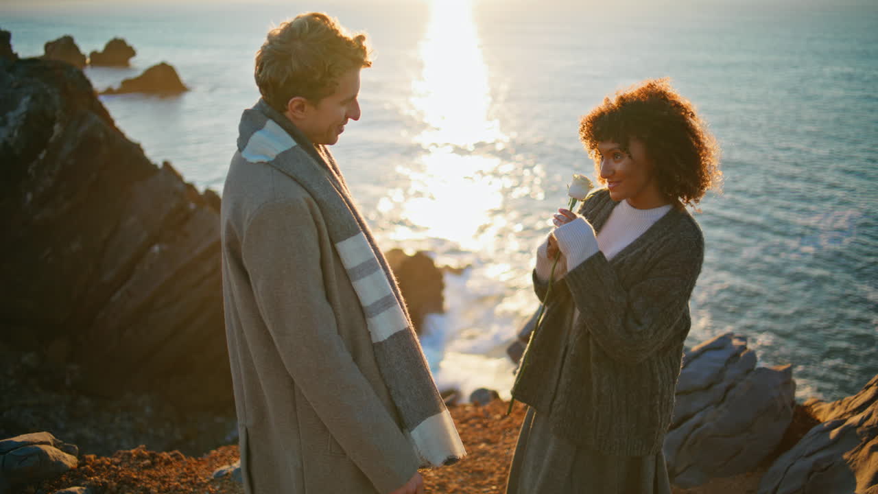 Lovely pair dating sunset sea cliffs closeup. Enamoured man bringing flower
