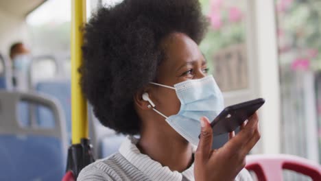 mujer de negocios afroamericana con máscara facial hablando en un teléfono inteligente y sentada en el autobús