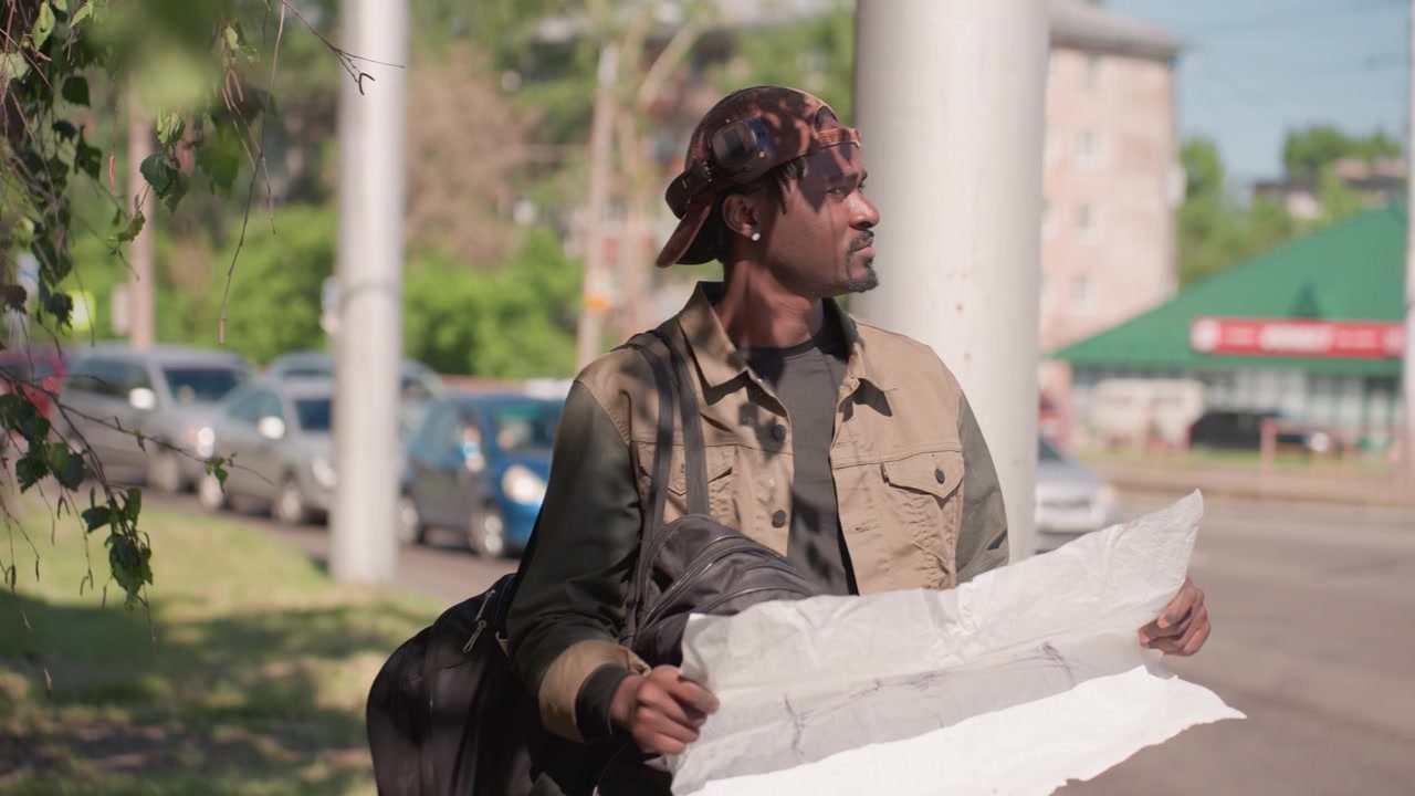 Un uomo esamina una grande mappa mentre è in piedi all'aperto vicino a auto parcheggiate e vegetazione, tenendo delle borse. L'uomo indossa abiti casual con protezione per le orecchie sul suo cappello, guardandosi intorno come se stesse navigando.