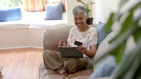 mujer biracial senior feliz en el sofá con tarjeta de crédito y usando tableta en casa, cámara lenta