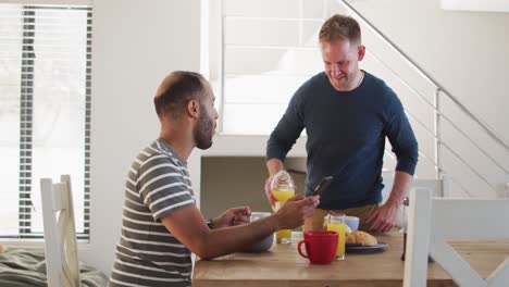 Multi-ethnic-gay-male-couple-having-breakfast-and-talking-one-using-smartphone