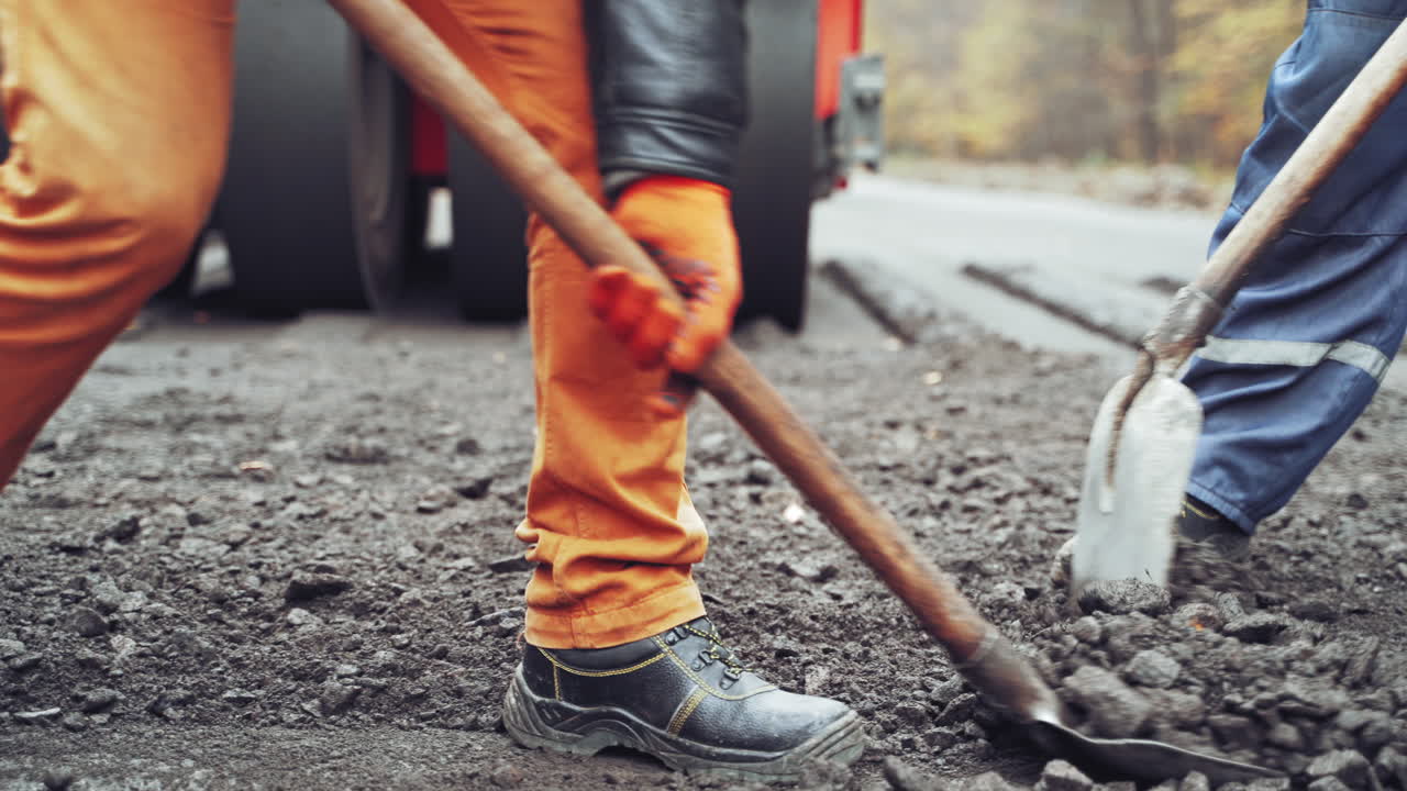 Team of workers put the hot asphalt on a street on the background of steamroller. Road construction workers with shovels in protective uniforms.