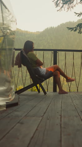 lonely woman rests in armchair on balcony overlooking forest and mountains with trees at sunset dusk in early evening. peace and relaxation in solitude