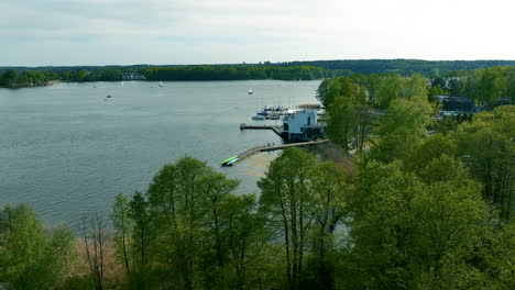 An-aerial-view-of-Ukiel-Lake-in-Olsztyn,-focusing-on-the-shoreline-with-boats-and-a-marina,-surrounded-by-green-trees-and-calm-water