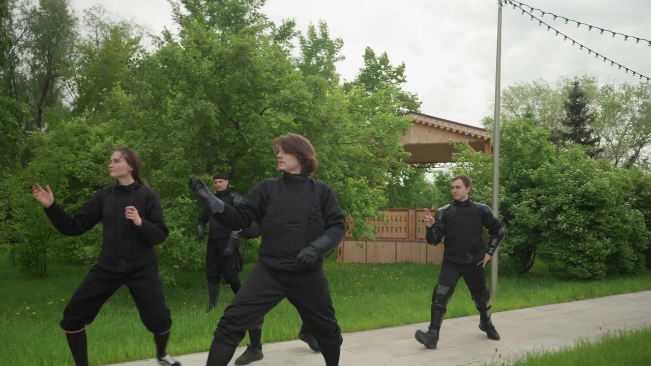 Gruppo di persone con equipaggiamento protettivo nero che praticano movimenti sincronizzati all'aperto su un sentiero pavimentato circondato da vegetazione e una struttura in legno. Le persone mantengono posizioni coordinate.
