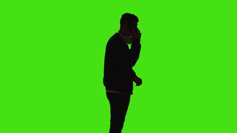 silhouette of man wearing headphones talking on mobile phone standing against green screen with low key lighting