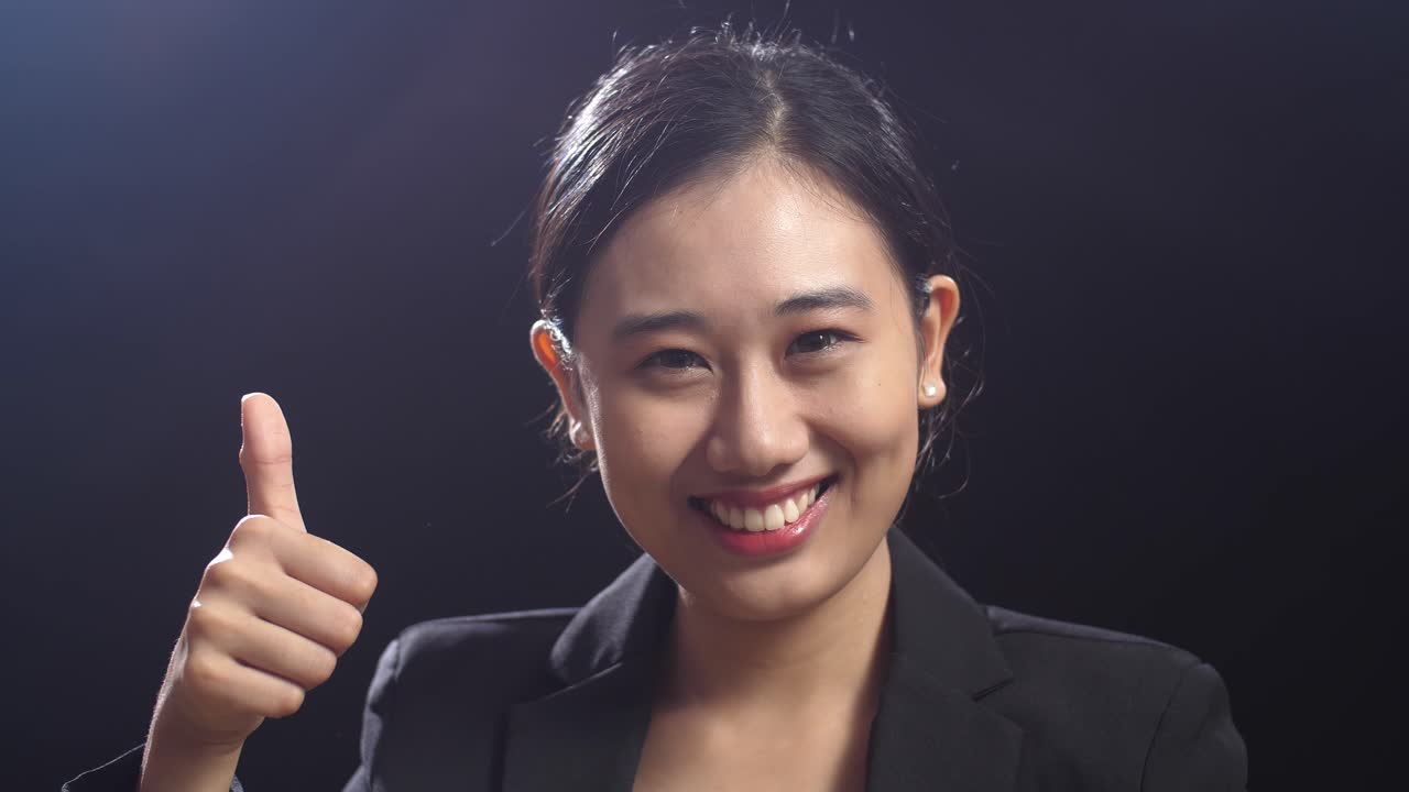 블랙 스크린 스튜디오에 서 있는 동안 엄지손가락을 올린 몸짓을 하는 비즈니스 슈트를 입은 미소 짓는 아시아 스피커 여성의 클로즈업