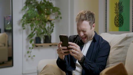 man browsing internet watching videos on social media and having fun with his smartphone comfortably, living room at home