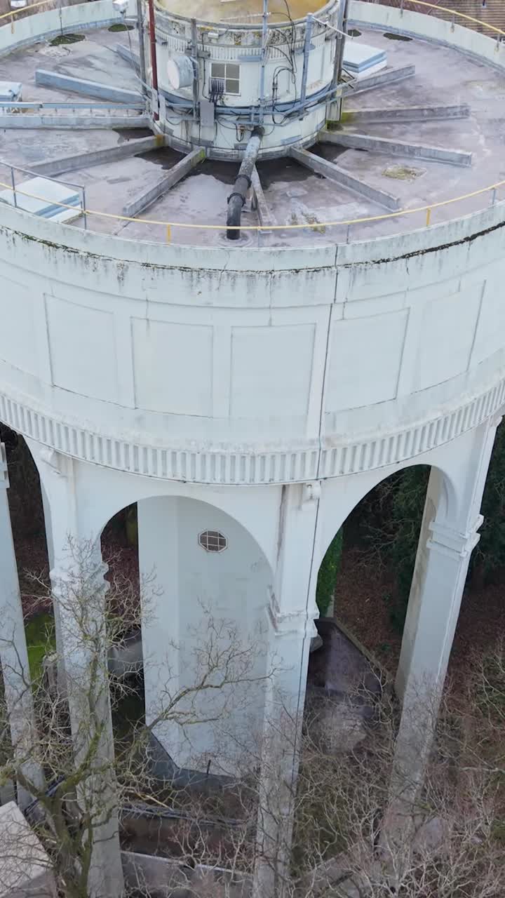 Ascesa verticale del drone alla torre dell'acqua di Bengeo con inquadratura fissa che rivela le antenne di comunicazione sui tetti, quindi si apre sui tetti dei villaggi dell'Hertfordshire, alberi e orizzonte della campagna